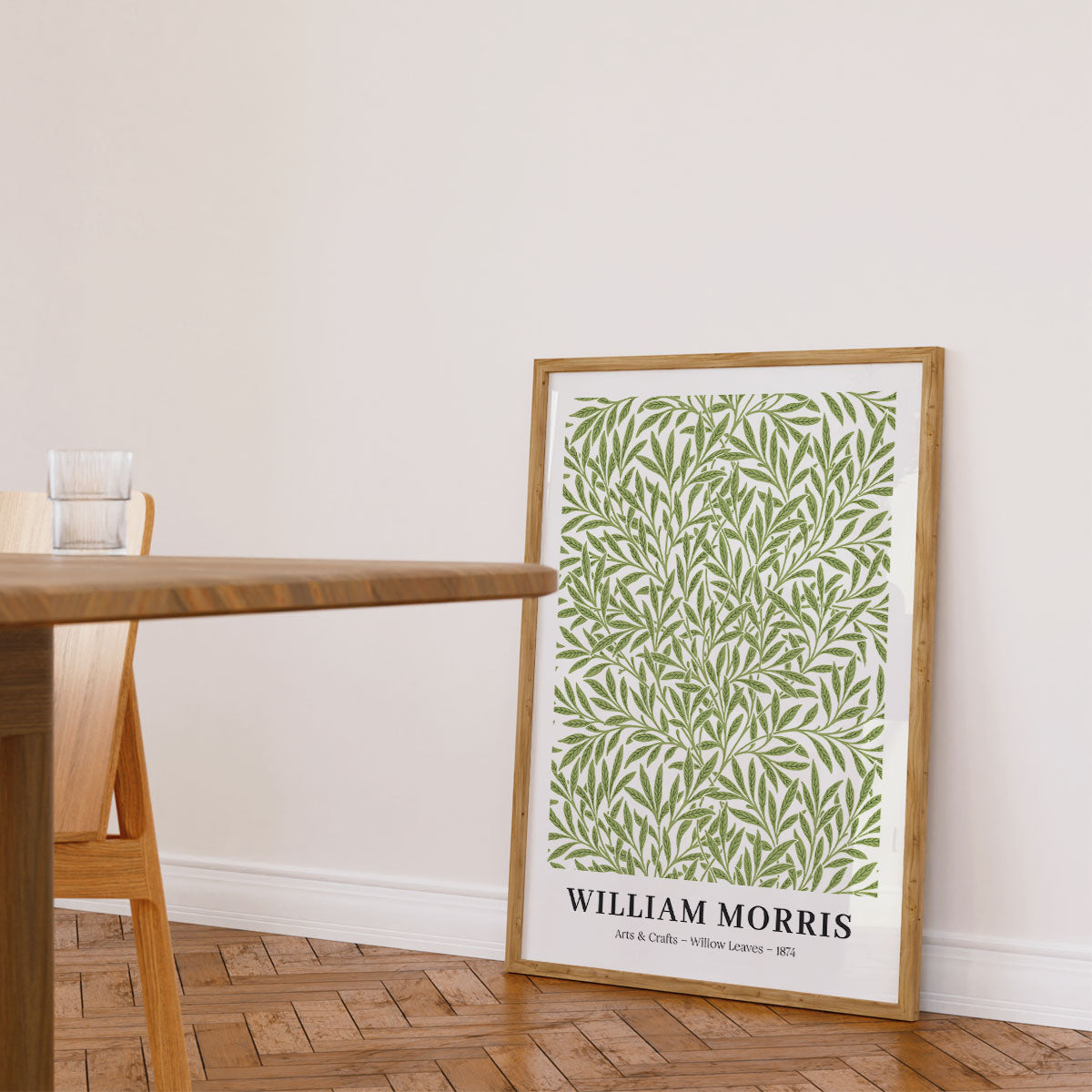 Plakat af William Morris med titlen "Willow Leaves" i en træramme, placeret på gulvet ved siden af et træbord og en stol i en lys stue.