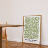 Plakat af William Morris med titlen "Willow Leaves" i en træramme, placeret på gulvet ved siden af et træbord og en stol i en lys stue.