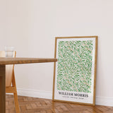 Plakat af William Morris med titlen "Willow Bough" i en træramme, placeret på gulvet i en stue ved siden af et træbord.