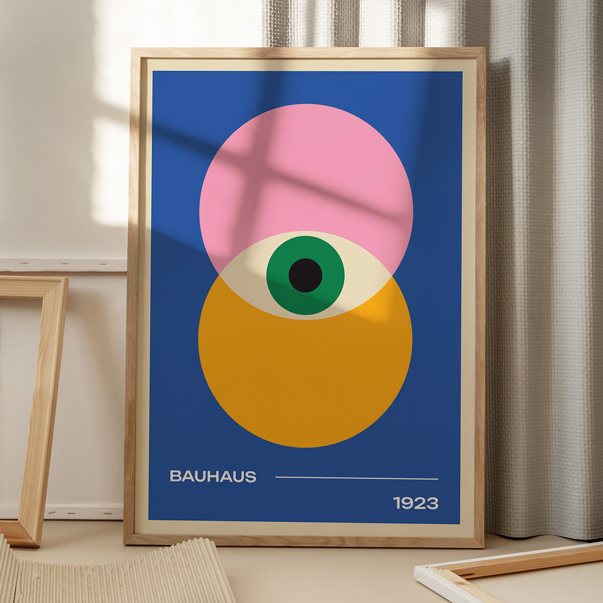 Plakat med Bauhaus-design fra 1923, viser geometriske former i pink, gul og grøn på en blå baggrund, placeret på et gulv med skygger fra et vindue.