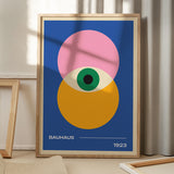 Plakat med Bauhaus-design fra 1923, viser geometriske former i pink, gul og grøn på en blå baggrund, placeret på et gulv med skygger fra et vindue.