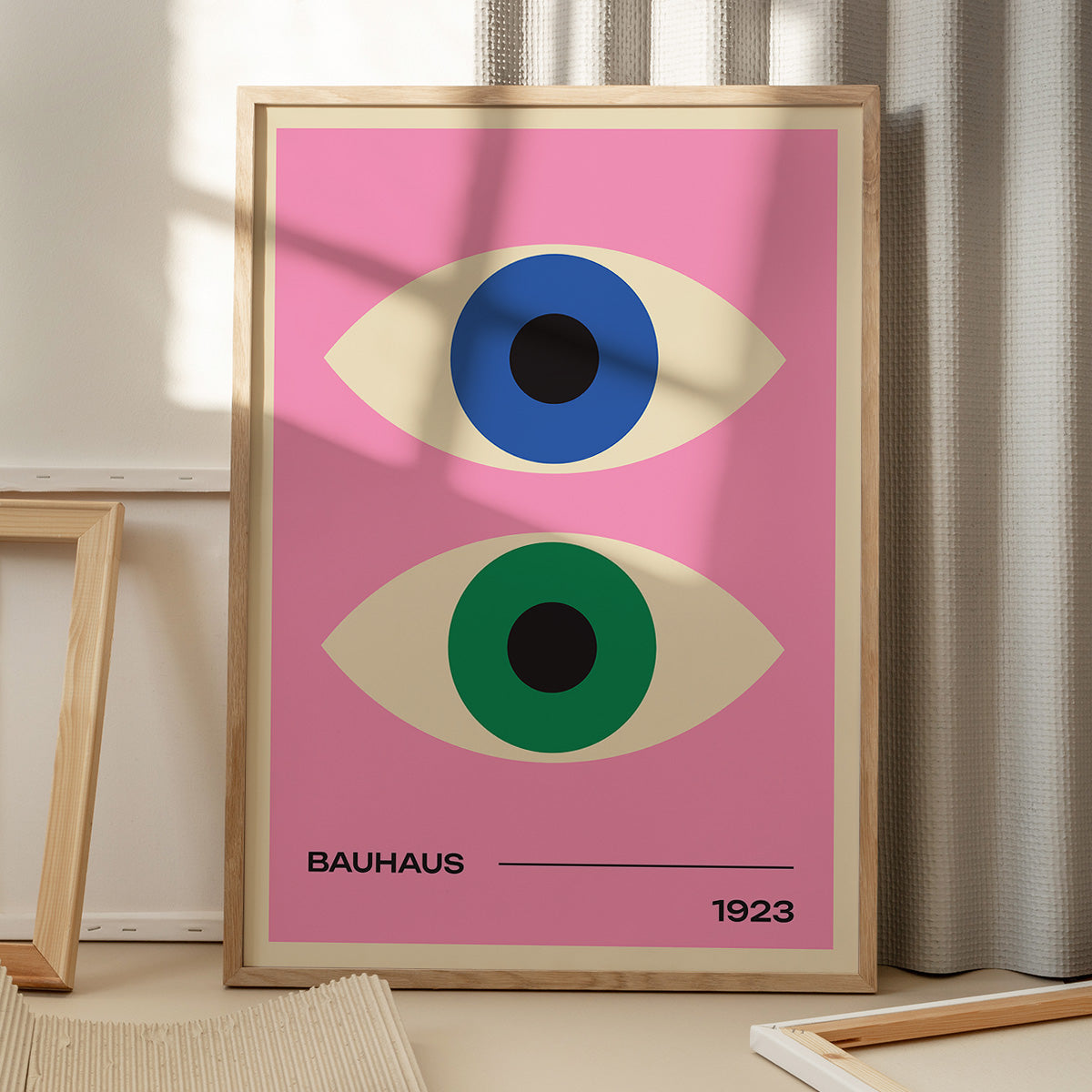 Plakat med to stiliserede øjne i blå og grøn på en lyserød baggrund, med teksten "Bauhaus 1923" i bunden, placeret på et gulv med skygger fra et vindue.