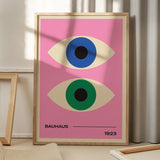 Plakat med to stiliserede øjne i blå og grøn på en lyserød baggrund, med teksten "Bauhaus 1923" i bunden, placeret på et gulv med skygger fra et vindue.