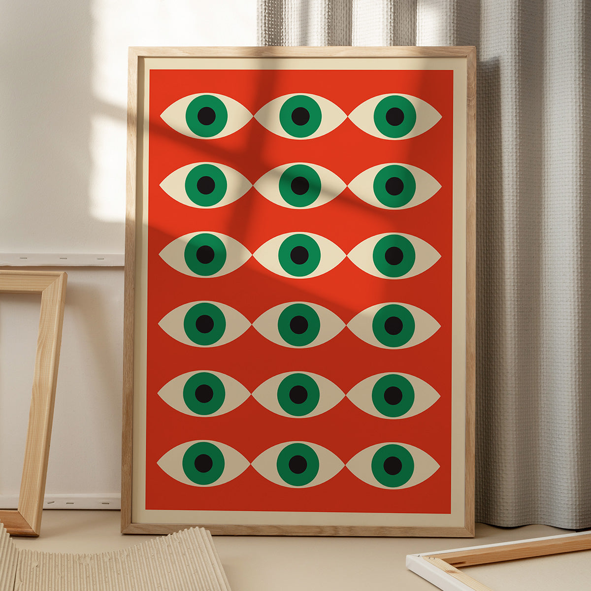 Plakat med et geometrisk øje-mønster i grøn og hvid på en rød baggrund, placeret på et gulv med skygger fra et vindue.