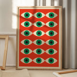 Plakat med et geometrisk øje-mønster i grøn og hvid på en rød baggrund, placeret på et gulv med skygger fra et vindue.