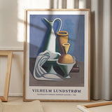 Plakat af Vilhelm Lundstrøm med titlen "Opstilling med vandkande, håndklæde og krukker - 1929" i en træramme, placeret på et gulv med skygger fra et vindue.