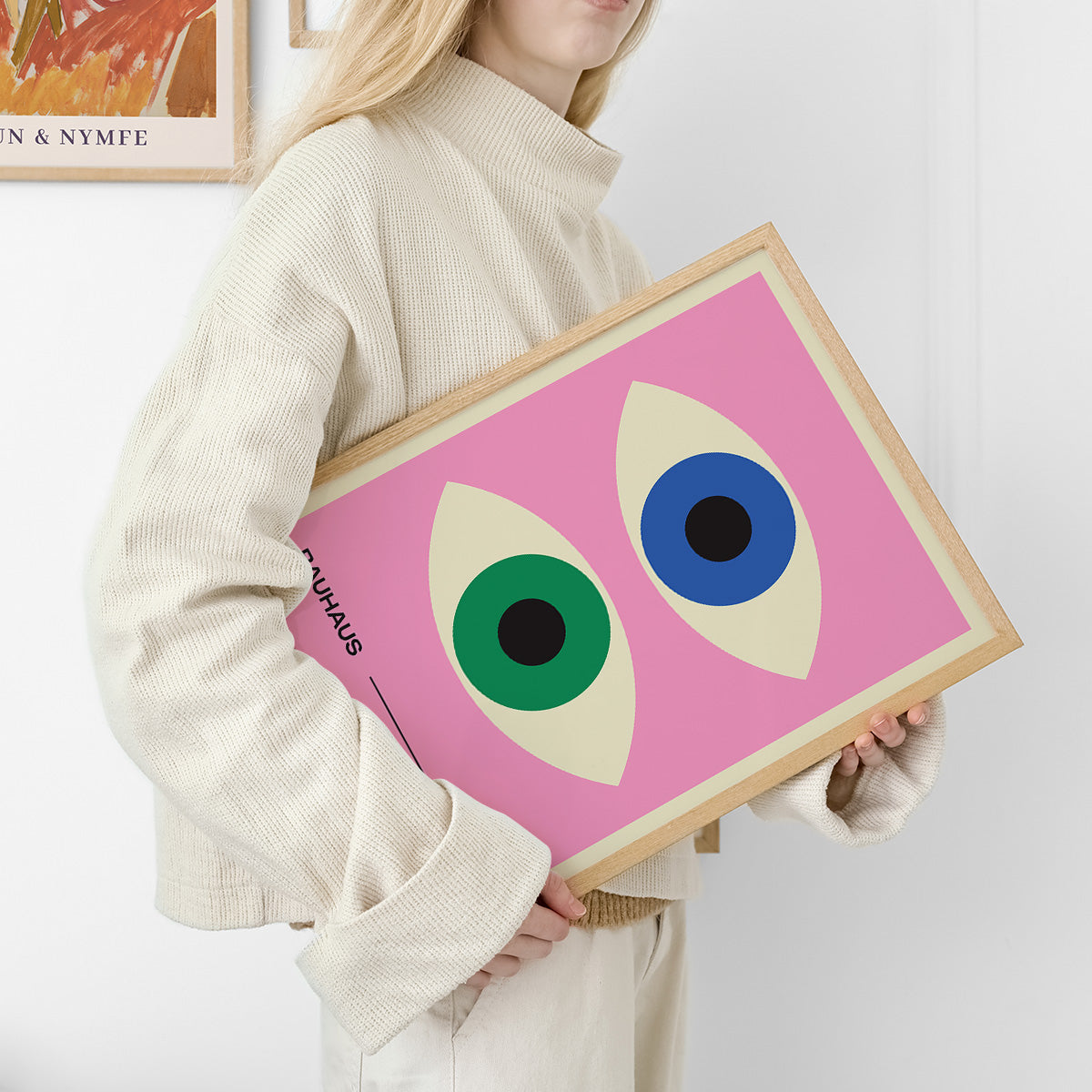 Kvinde holder en Bauhaus-plakat med et pink baggrund og to øjenlignende figurer i grøn og blå.