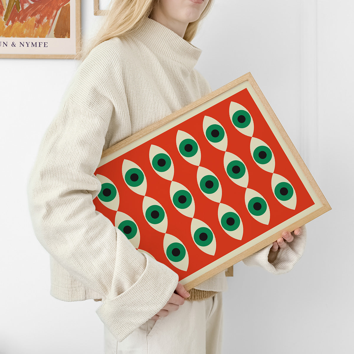Plakat med geometrisk mønster af grønne øjne på rød baggrund, holdt af person i lys sweater.