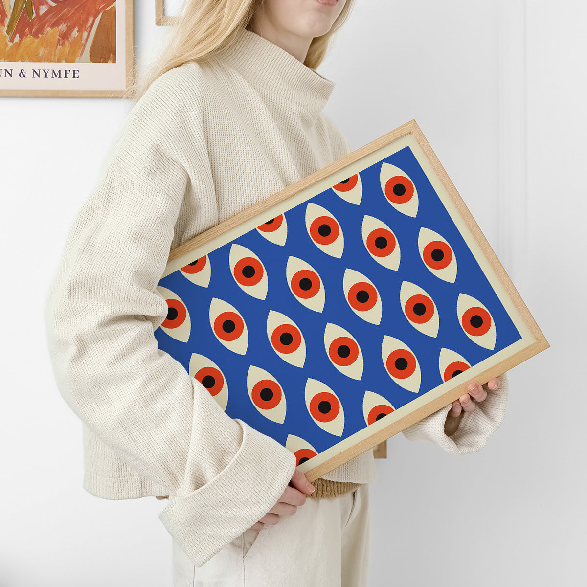 Kvinde i lys sweater holder en plakat med et geometrisk mønster af øjeformer i blå, rød og beige farver.