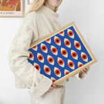 Kvinde i lys sweater holder en plakat med et geometrisk mønster af øjeformer i blå, rød og beige farver.