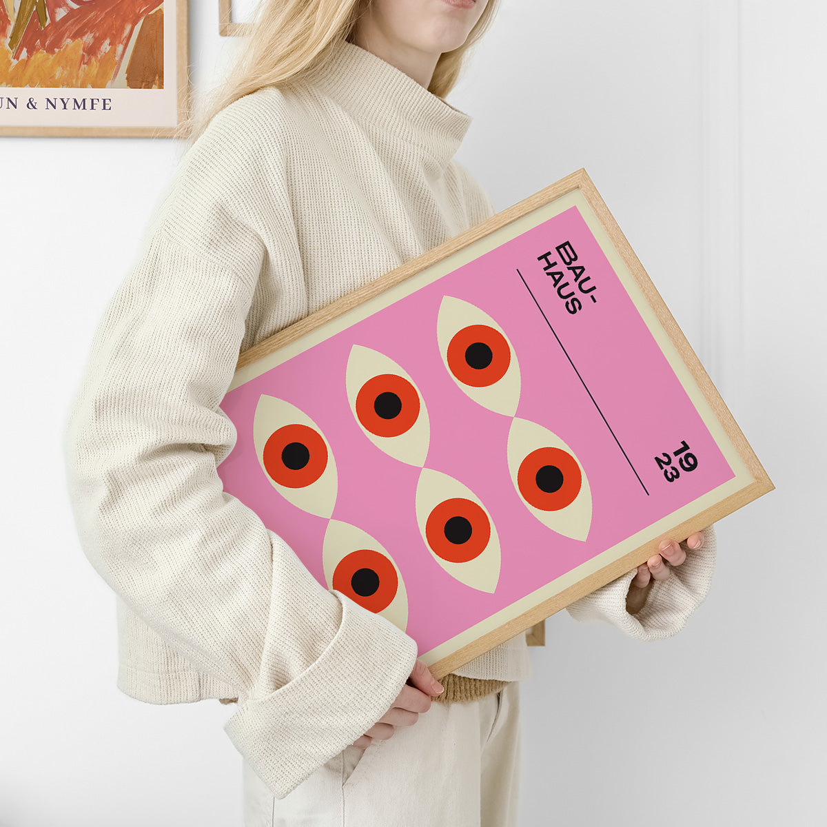 Kvinde holder en Bauhaus-plakat med et geometrisk design af øjenlignende figurer på en lyserød baggrund.