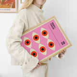 Kvinde holder en Bauhaus-plakat med et geometrisk design af øjenlignende figurer på en lyserød baggrund.