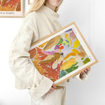 Kvinde i lys sweater holder en farverig plakat med abstrakt kunst i træramme.