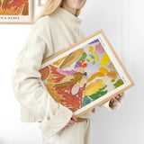 Kvinde i lys sweater holder en farverig plakat med abstrakt kunst i træramme.