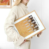 Kvinde i beige sweater holder en indrammet plakat af Gustav Klimt med et abstrakt design.