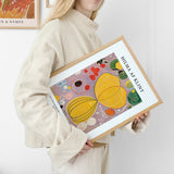 Kvinde holder en indrammet plakat af Hilma af Klint med abstrakt kunst i gule og lilla nuancer.