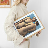 Kvinde i beige sweater holder en indrammet plakat med et kunstværk af to figurer.