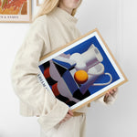 Kvinde i beige sweater holder en indrammet plakat med et abstrakt motiv af kander og en appelsin, signeret Vilhelm Lunds.