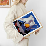 Kvinde i beige sweater holder en indrammet plakat med et abstrakt motiv af kander og en appelsin, signeret Vilhelm Lunds.