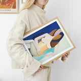 Kvinde i beige sweater holder en indrammet plakat med abstrakt kunst i blå, beige og brune nuancer.