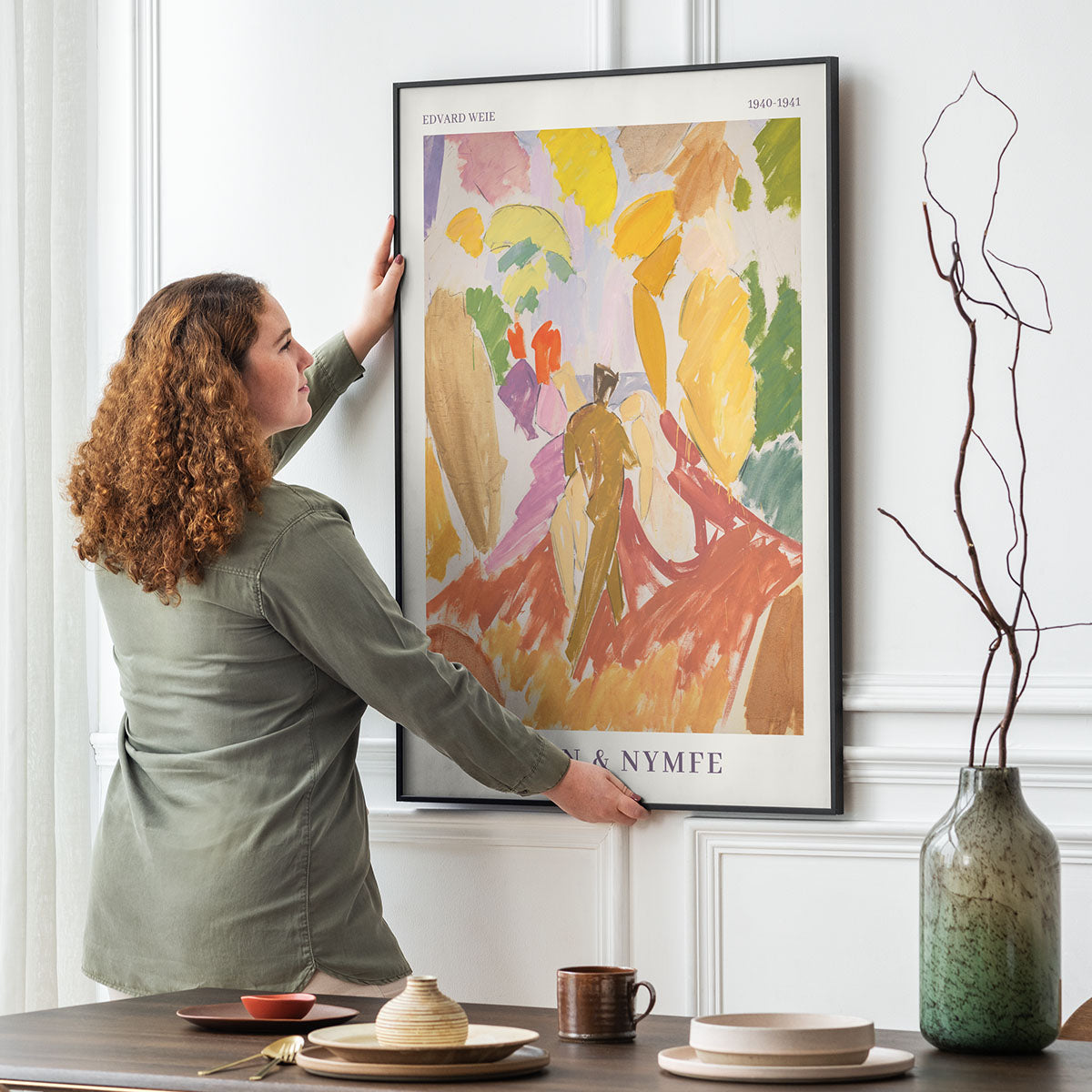 Kvinde hænger Faun og Nymfe kunstplakat af Edvard Weie op i en spisestue.
