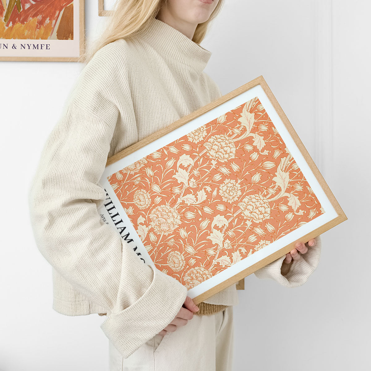 Kvinde i lys sweater holder en indrammet plakat med et blomstermønster i orange og beige, designet af William Morris.